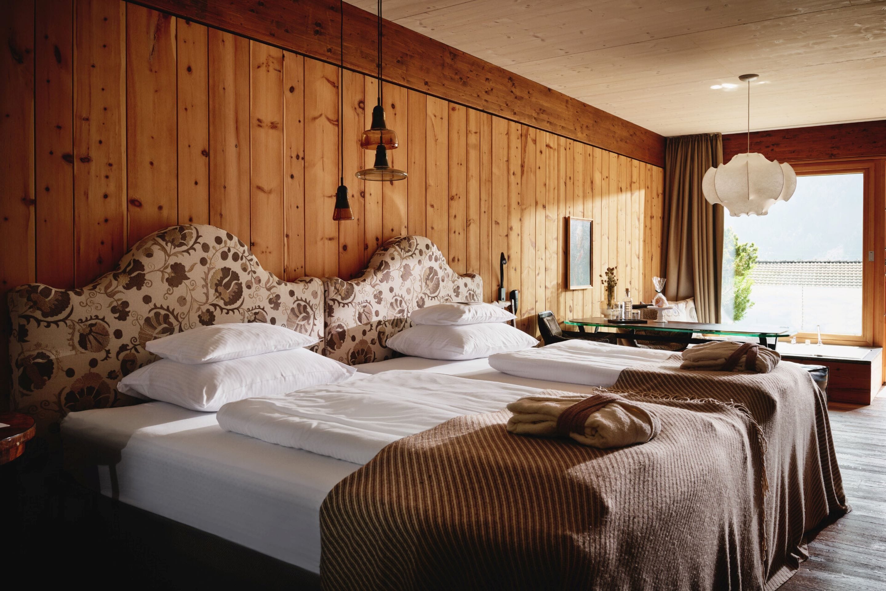 A hotel room with a wooden paneled wall, a large bed with patterned bedding, a bedside table, and a window with a view.