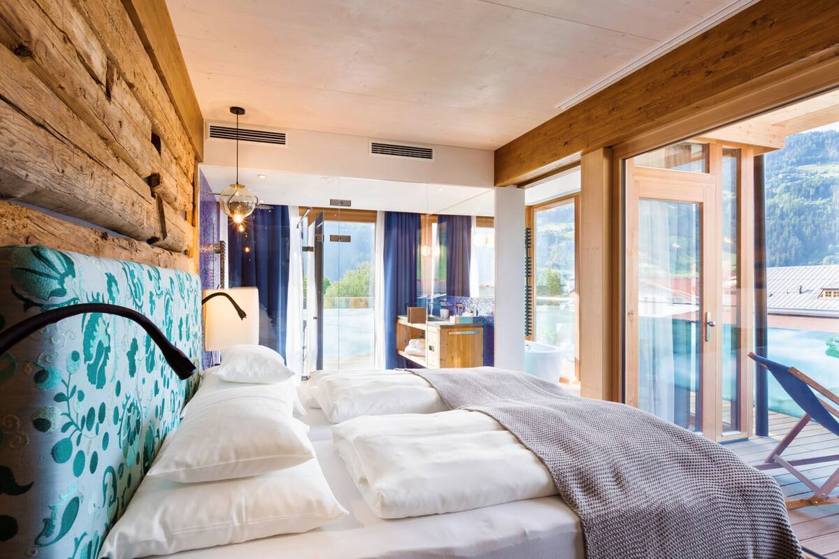Une chambre à coucher avec une tête de lit en bois, un grand lit recouvert de draps blancs, une vue sur un balcon et un plafond en bois.