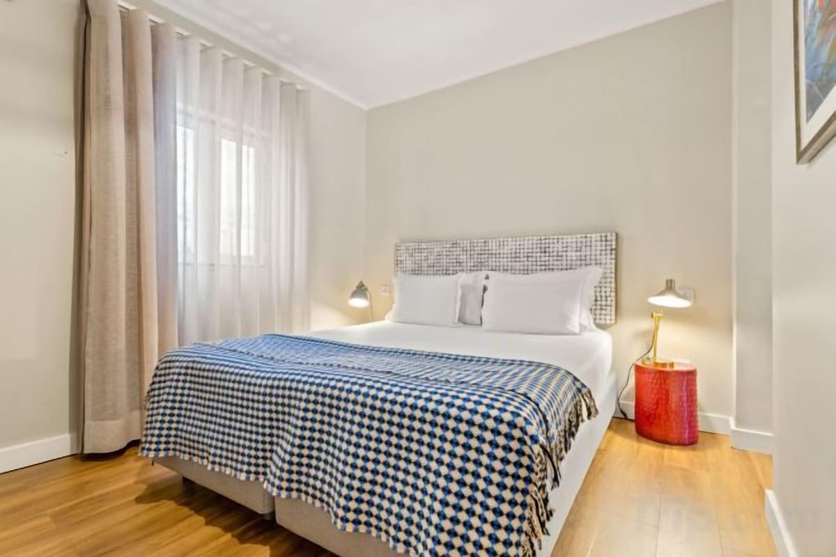 Ein Schlafzimmer mit einem Bett, zwei Nachttischlampen, einem Fenster mit Vorhängen und einem Holzboden.