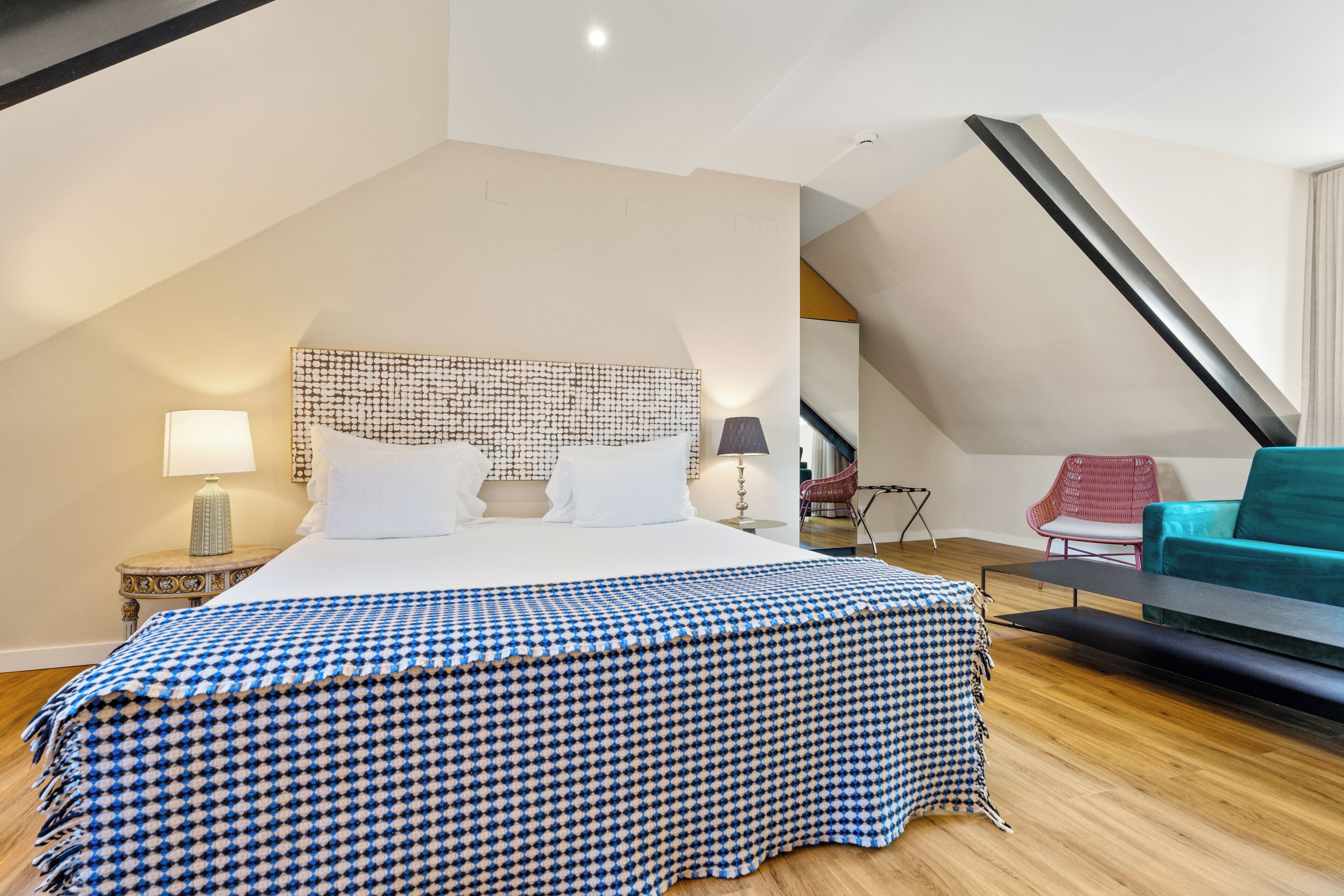 Un dormitorio con una cama grande, una colcha de cuadros de piedras, un cabecero con diseño geométrico, una mesita de noche pequeña con una lámpara y una silla roja.