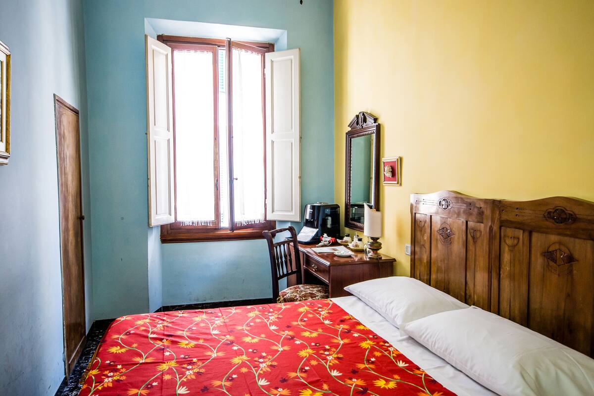 A bedroom with a bed, a desk with a computer, a chair, and a window with open shutters.