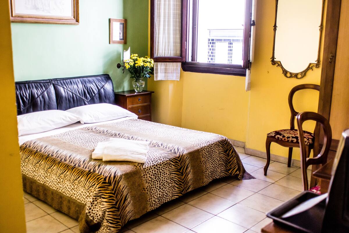 A bedroom with a zebra-patterned bedspread, a wooden nightstand with a vase of flowers, a chair, and a window with curtains.