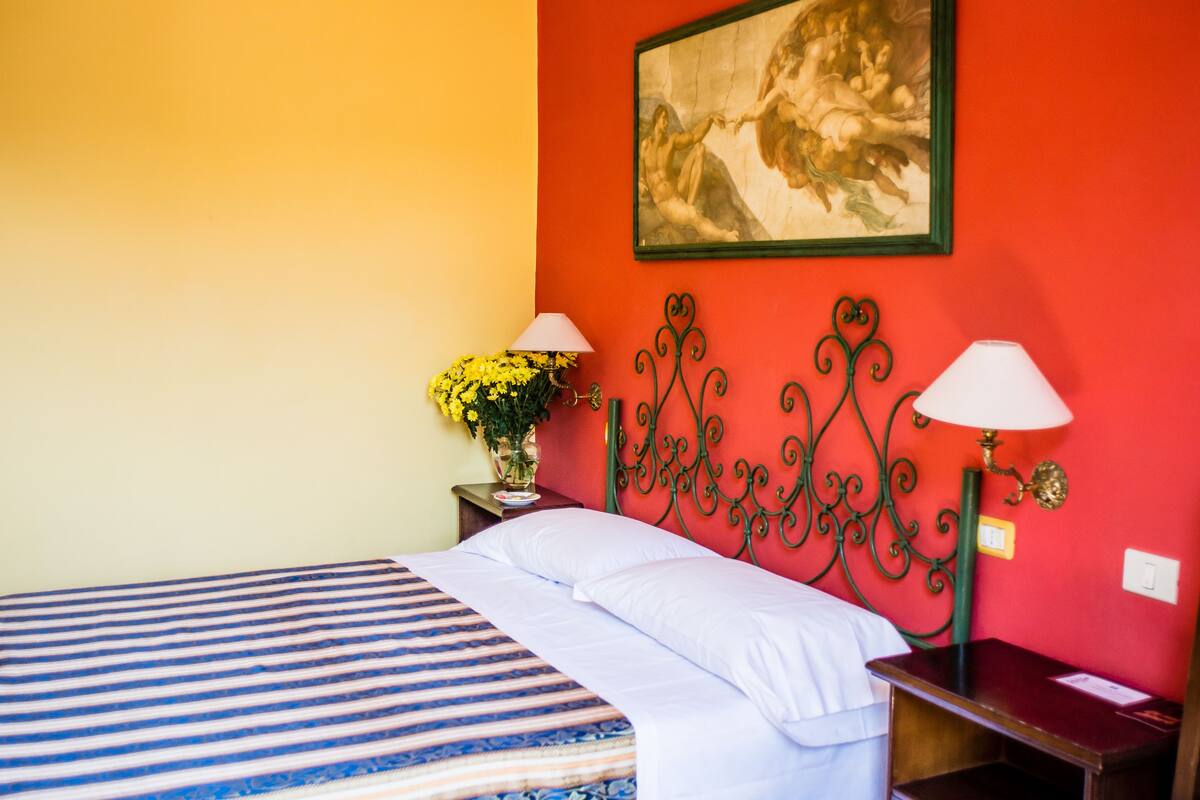 A hotel room with a bed, a nightstand with a lamp, a vase with flowers, and a painting on the wall.