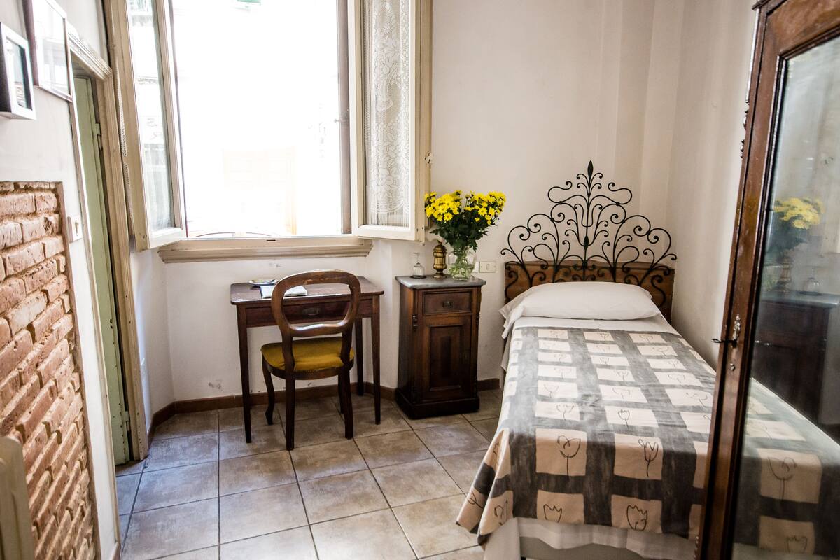 A bedroom with a bed, a desk, a chair, a small table with flowers, and a window.