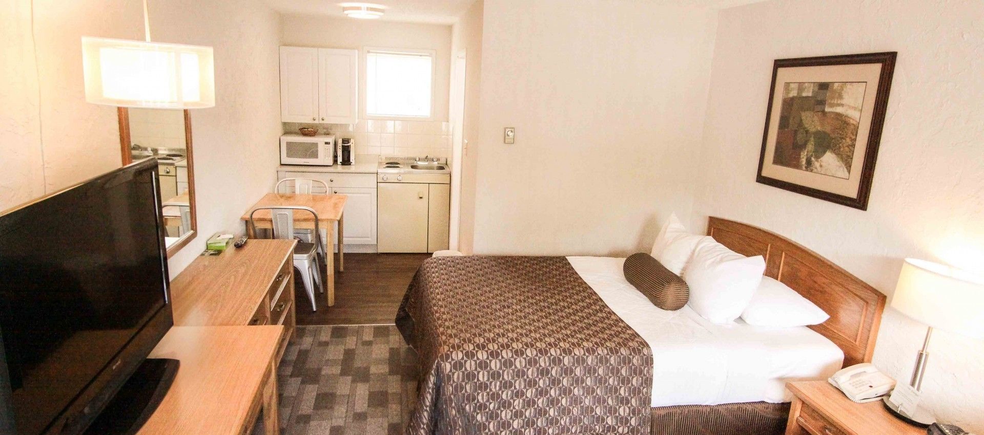 A hotel room with a bed, bedside table, TV, kitchenette, and a framed picture on the wall.