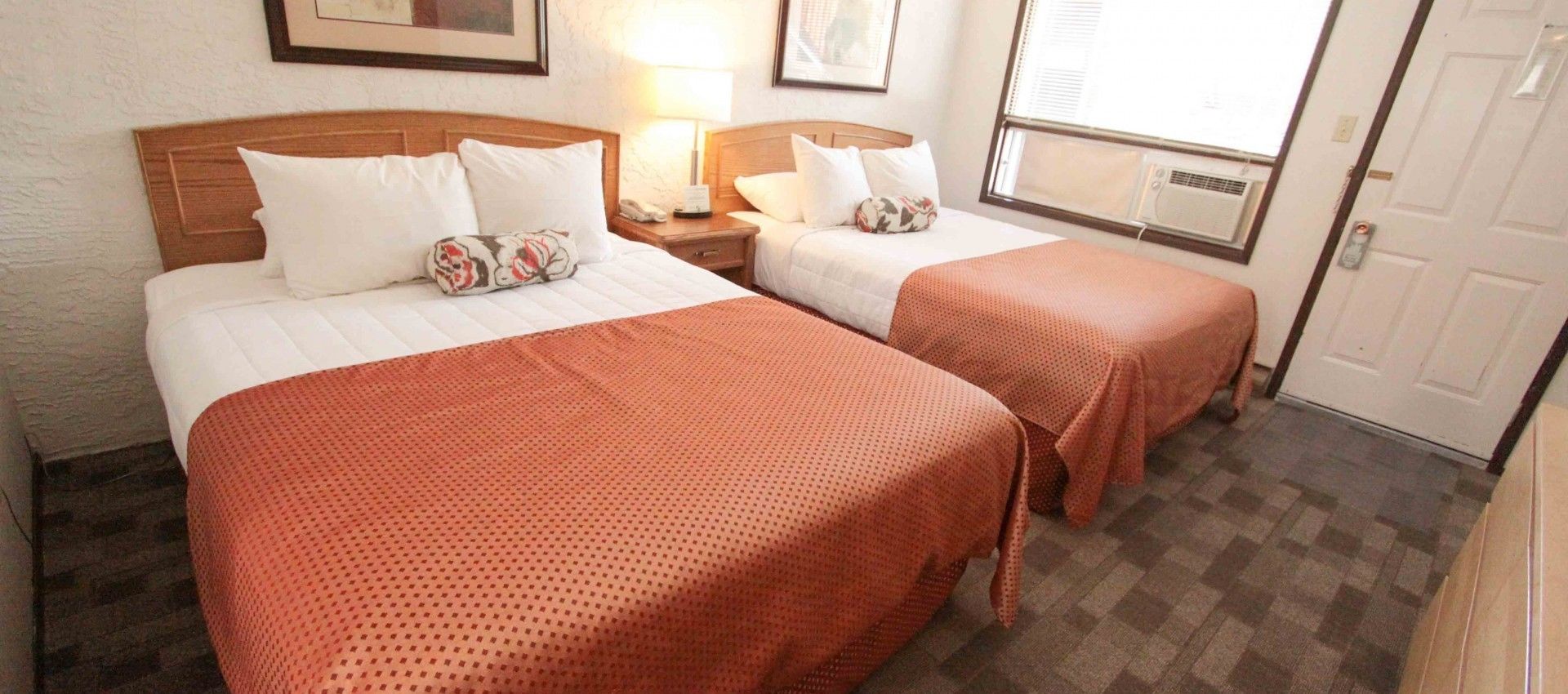 A hotel room with two beds, each with a wooden headboard, white bedding, and a patterned pillow. There is a nightstand with a lamp, a window with blinds, and a door.