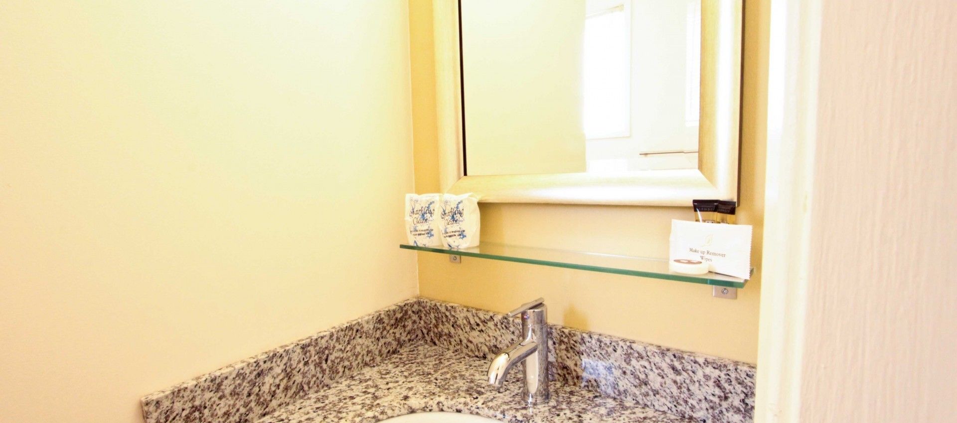 A bathroom with a granite countertop, a sink, a mirror, and a shelf with toiletries.