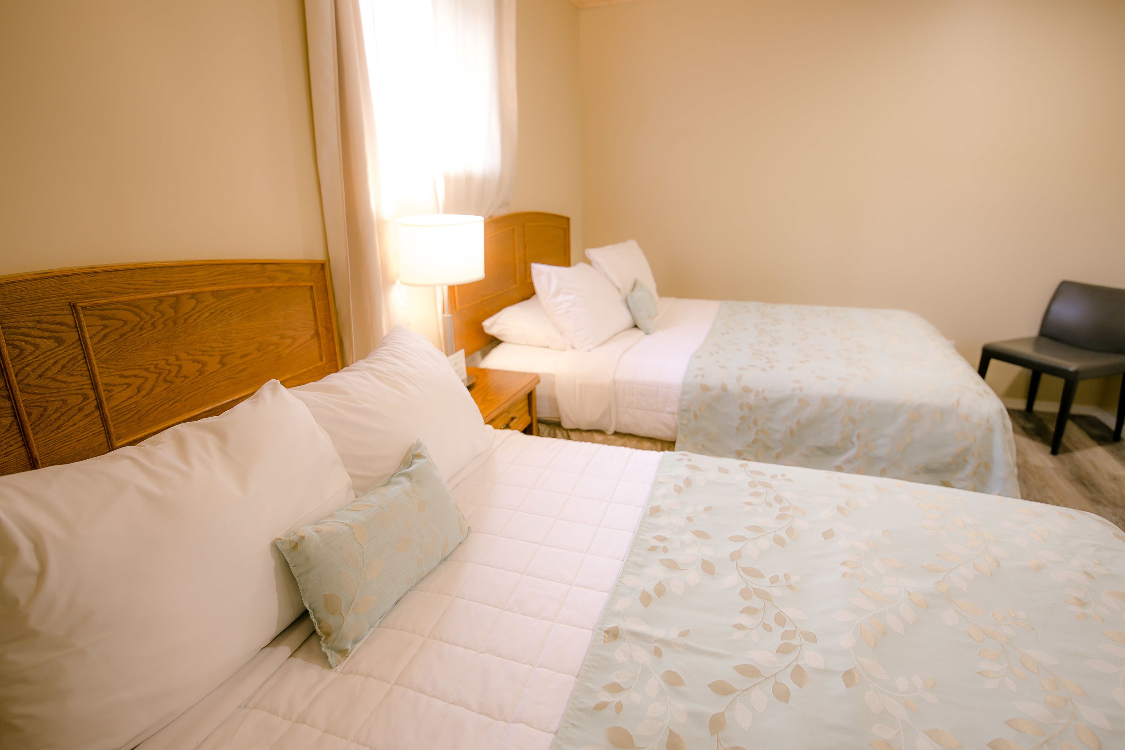 A hotel room with two beds, a nightstand, a lamp, and a chair.