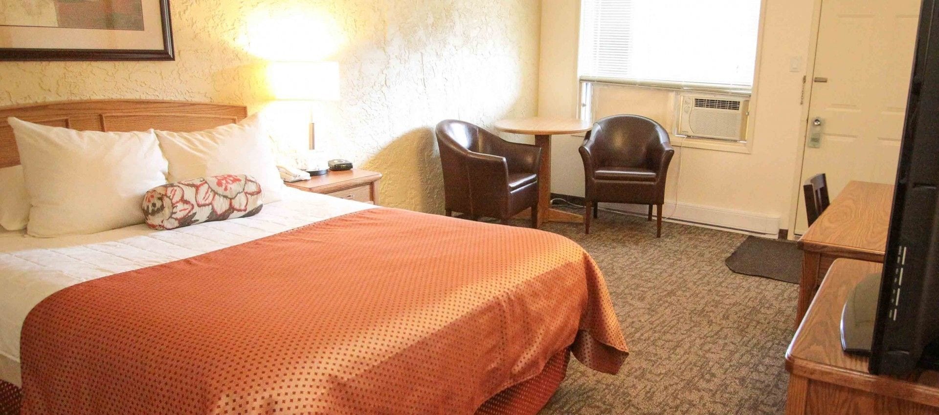Habitación de hotel con cama, mesita de noche, escritorio, dos sillas, lámpara y ventana con persianas.