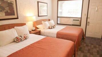 Habitación de hotel con dos camas, cada una con cabecero de madera, ropa de cama blanca y una manta roja. Se ve una ventana con persianas, una puerta y un cuadro enmarcado en la pared.