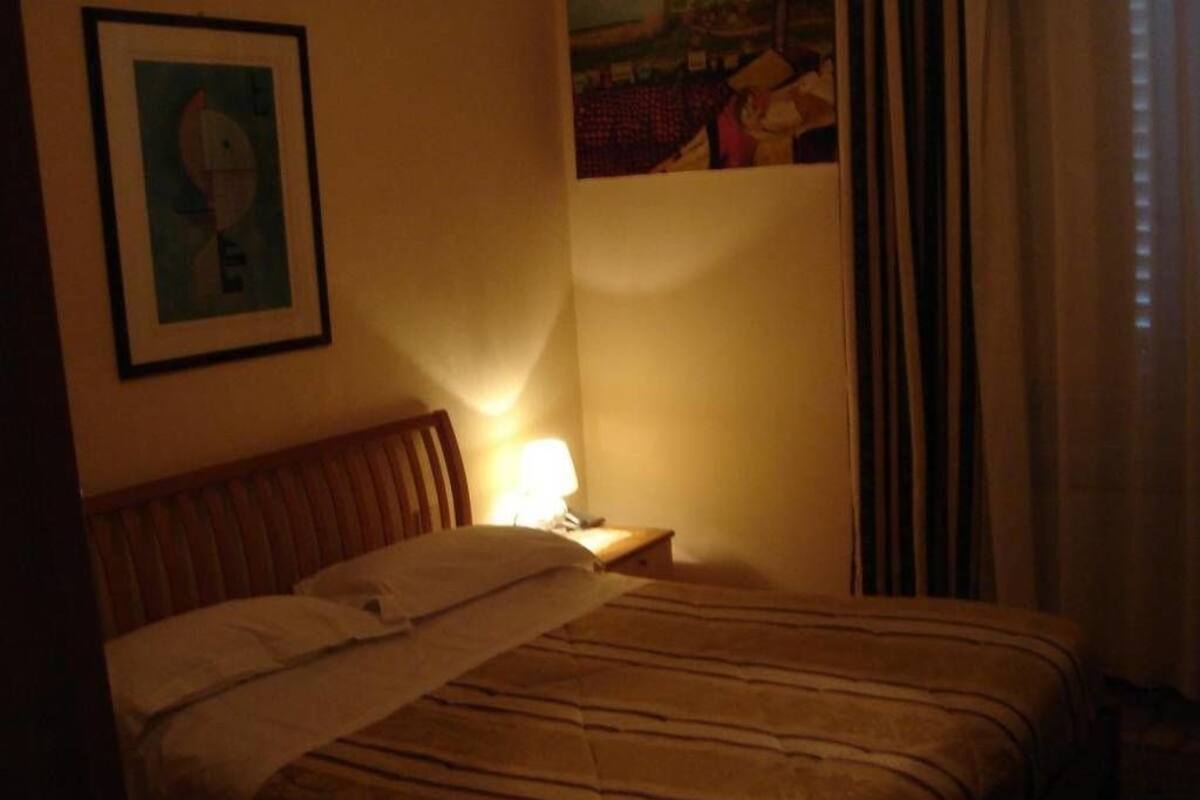 A dimly lit hotel room with a bed, a nightstand, a lamp, and a framed picture on the wall.