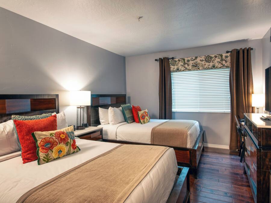 A hotel room with two beds, a desk, a lamp, and a TV.