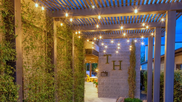 A covered walkway with string lights, a stone wall with the letter "H", and a clear path leading to a building entrance.