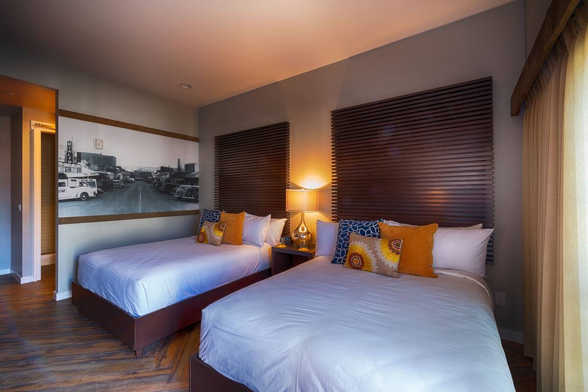 Ein Hotelzimmer mit zwei Betten, Holz-Kopfteilen, einem Stadtbild-Wandgemälde und einem Nachttisch mit Lampe.