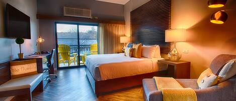 A modern hotel room with a large bed, a sofa, a desk, and a balcony with yellow chairs.