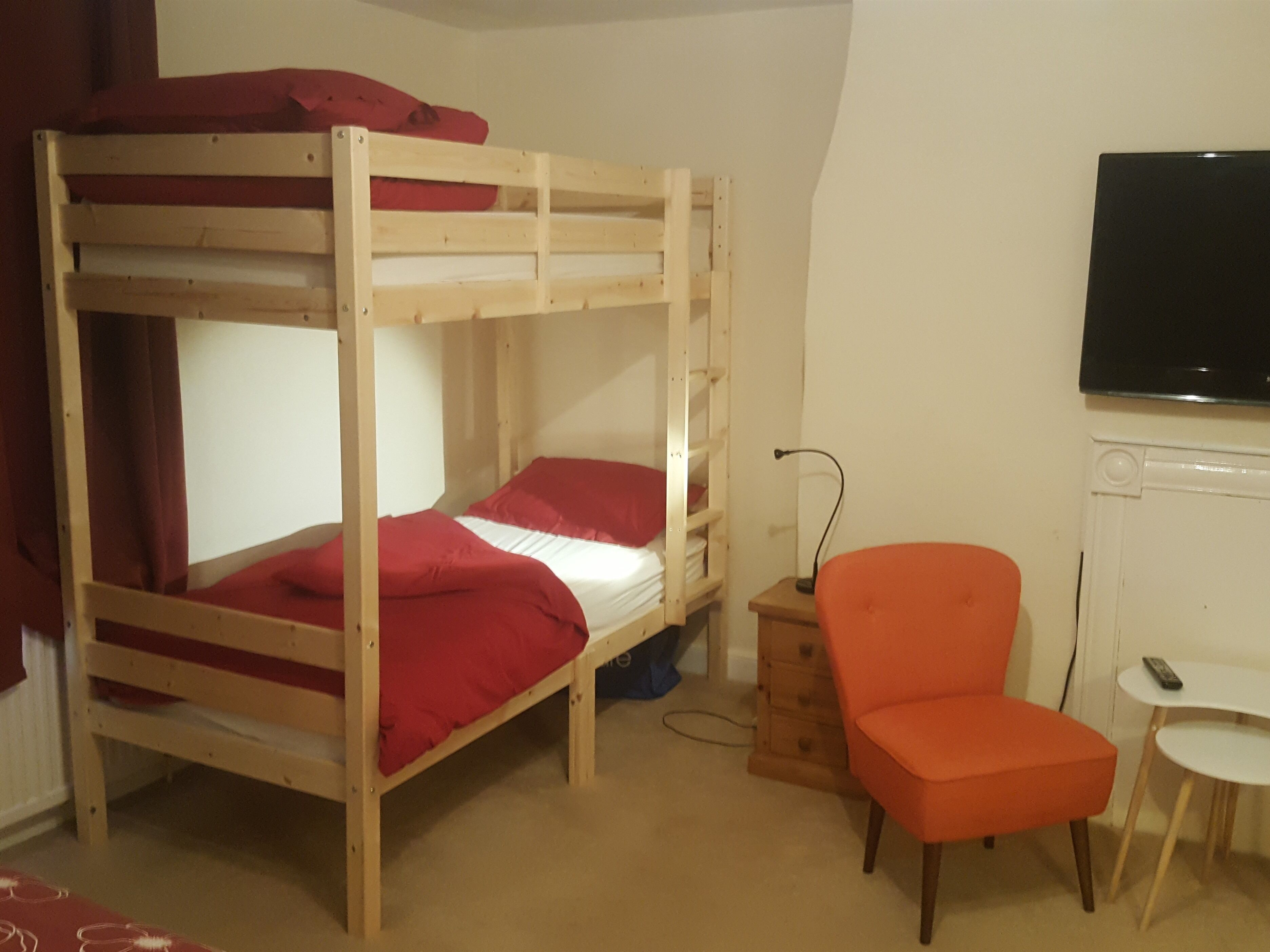 Una cama litera con ropa de cama roja, una mesita de noche de madera, una silla naranja y una pequeña mesa blanca con un control remoto.