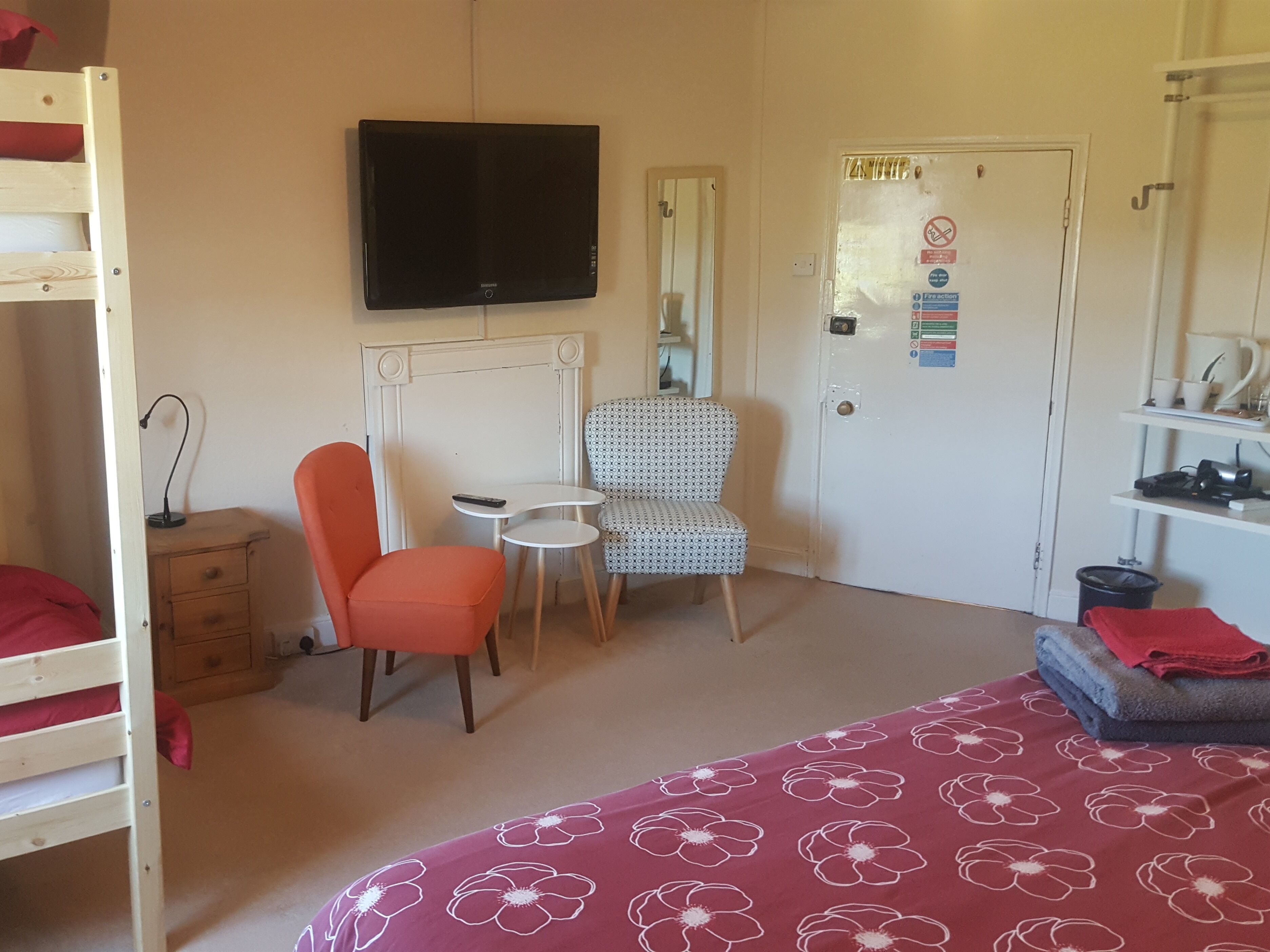 A room with a bunk bed, a single bed with a floral bedspread, a television, a chair, a small table, and a door with a fire safety sign.