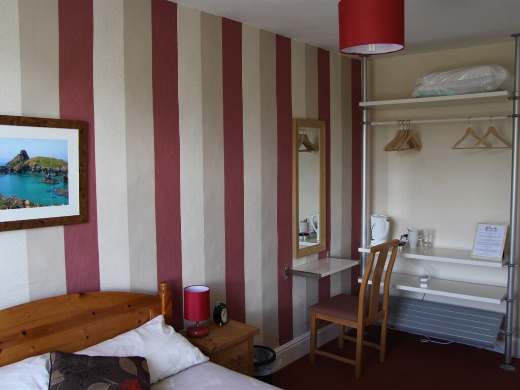 A hotel room with striped wallpaper, a bed with a wooden headboard, a desk with a chair, a mirror, a shelf with hangers, and a radiator.