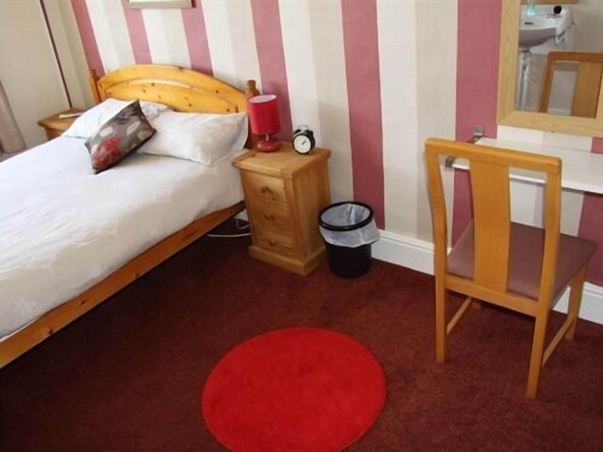 Une chambre à coucher avec un lit, une table de chevet en bois, une poubelle rouge, une chaise en bois et un miroir accroché au mur.