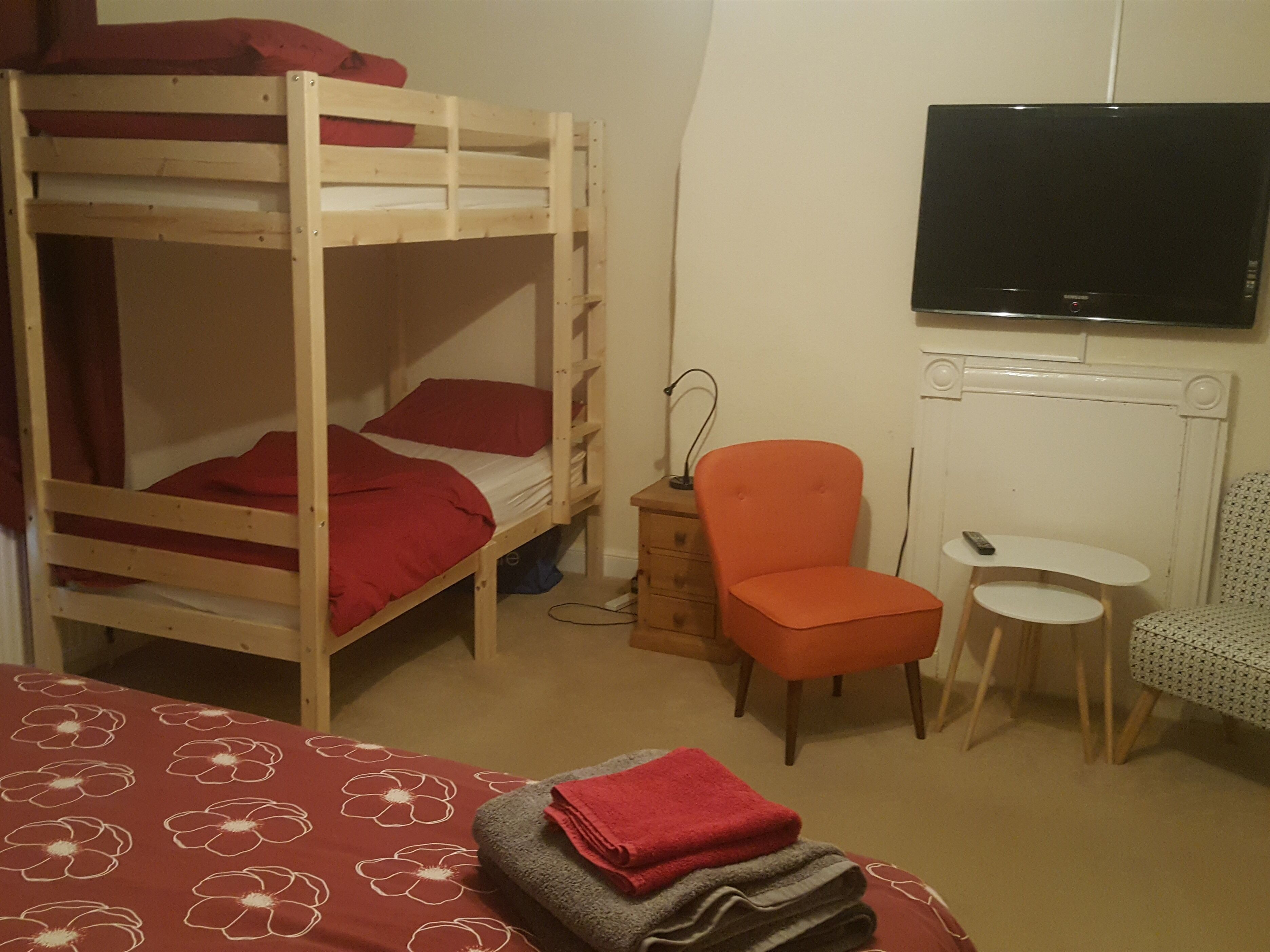 Una cama litera con ropa de cama roja, una pequeña silla naranja, una mesa blanca y un televisor en la pared.