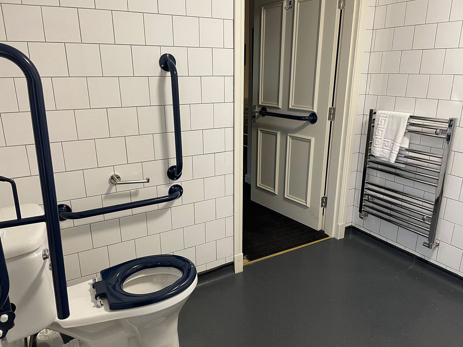 A bathroom with a white toilet, grab bars, a towel rack, and a door leading to another room.