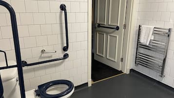Ein Badezimmer mit Toilette, Haltegriffen, einem Handtuchhalter und einer Tür zu einem anderen Raum.