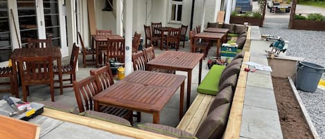 An outdoor patio area with wooden tables and chairs, a built-in bench with cushions, and umbrellas providing shade.
