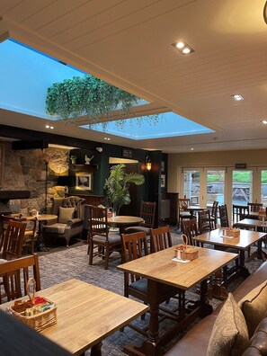 A spacious indoor area with multiple wooden tables and chairs, a stone wall, and a skylight with hanging plants. - The Bridge Inn (Holmrook)