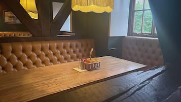 Un banco de madera con respaldo acolchado, una mesa de madera y una cesta de aperitivos, y una ventana con cortinas.