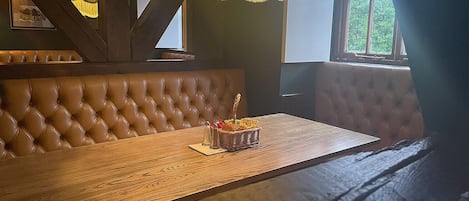 En träbänk med ett vadderat ryggstöd, ett träbord med en korg full av snacks och ett fönster med gardiner.