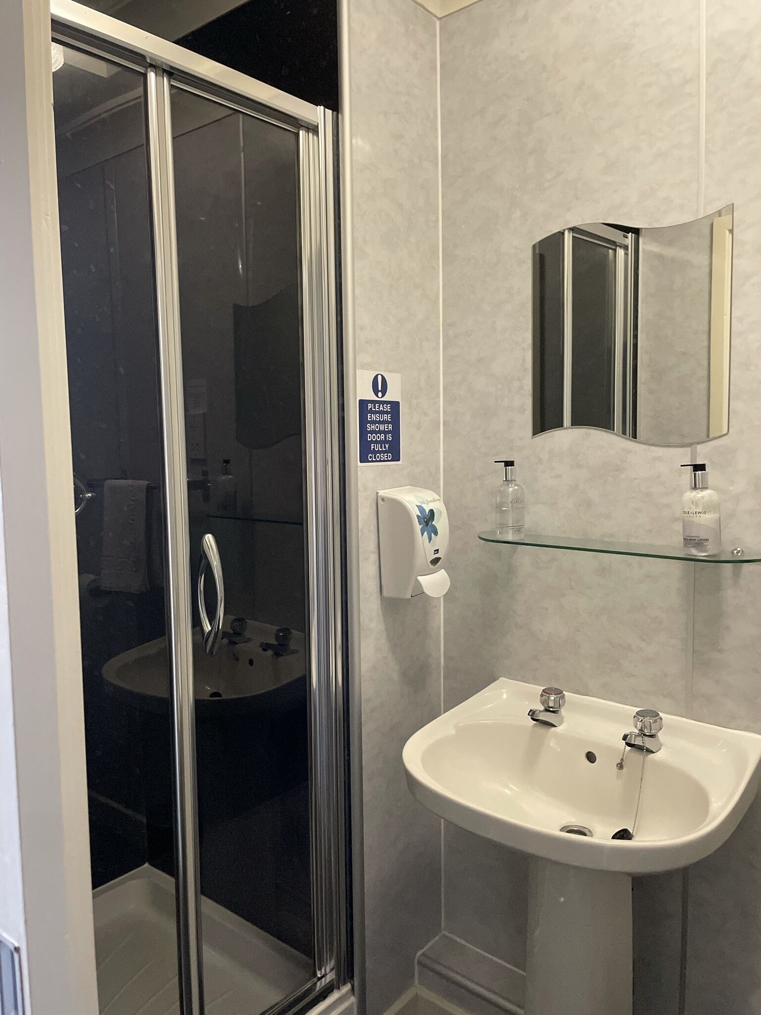 A modern bathroom with a glass shower door, a sink with two faucets, a mirror, and a soap dispenser on the wall.
