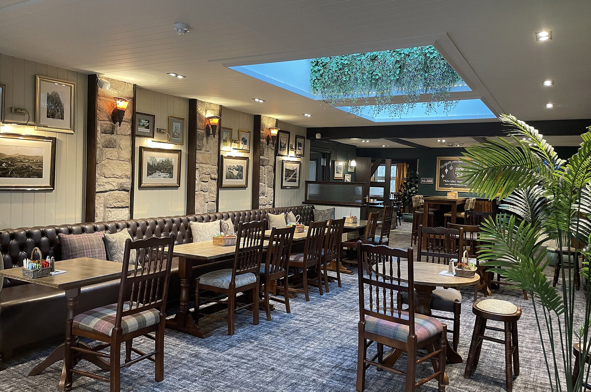 A restaurant with a high ceiling, stone walls, and framed pictures. There is a skylight and a large plant in the corner.