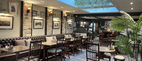 Un restaurante con techo alto, paredes de piedra y cuadros enmarcados. Hay un tragaluz y una planta grande en la esquina.