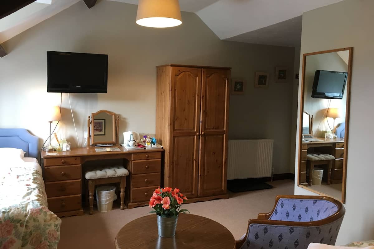 A bedroom with a wooden wardrobe, a desk with a lamp, a chair, a bed with a floral bedspread, and a television mounted on the wall.