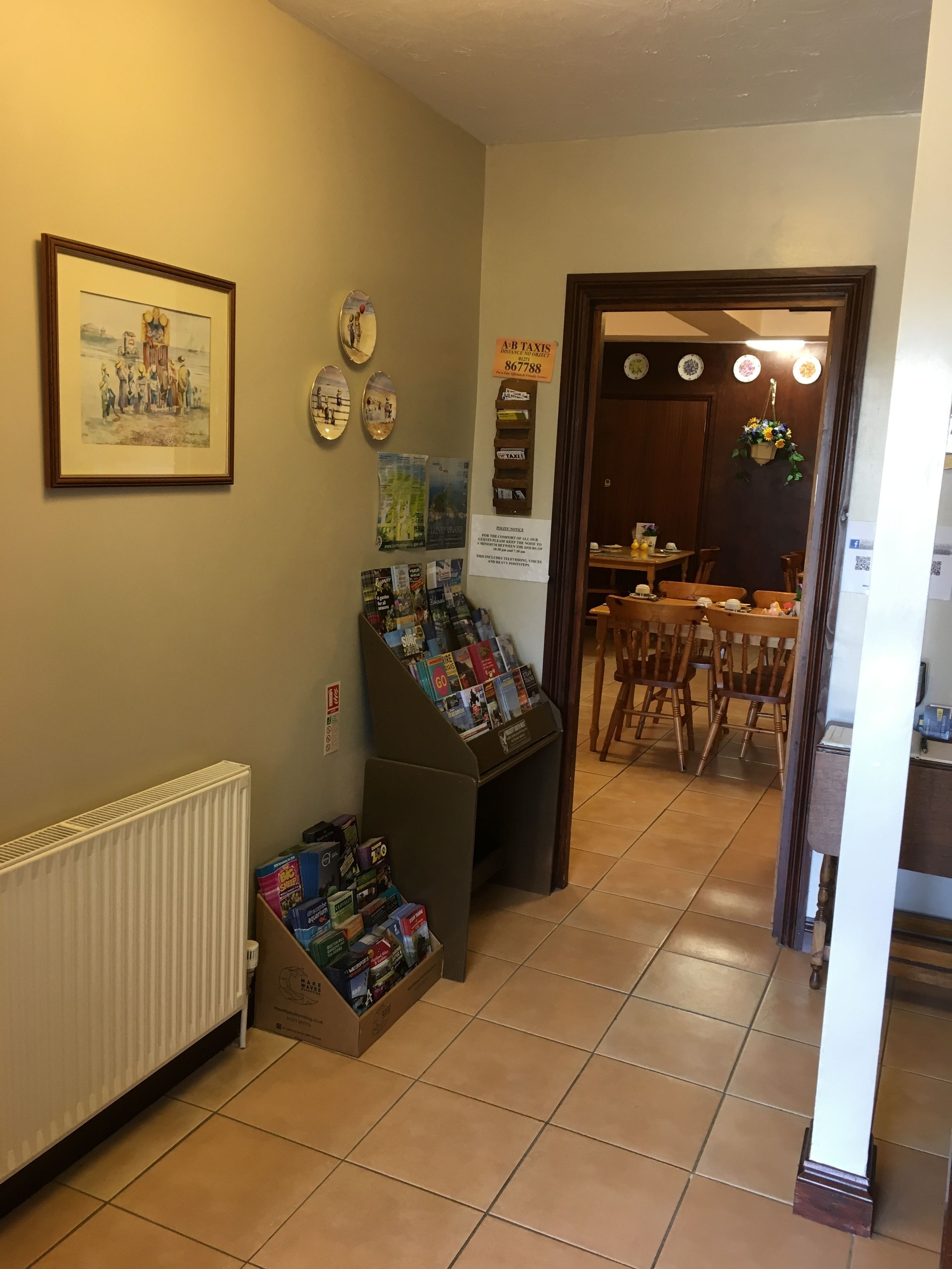 A hallway with a radiator, a display rack with magazines, and a dining area with wooden chairs and a table.