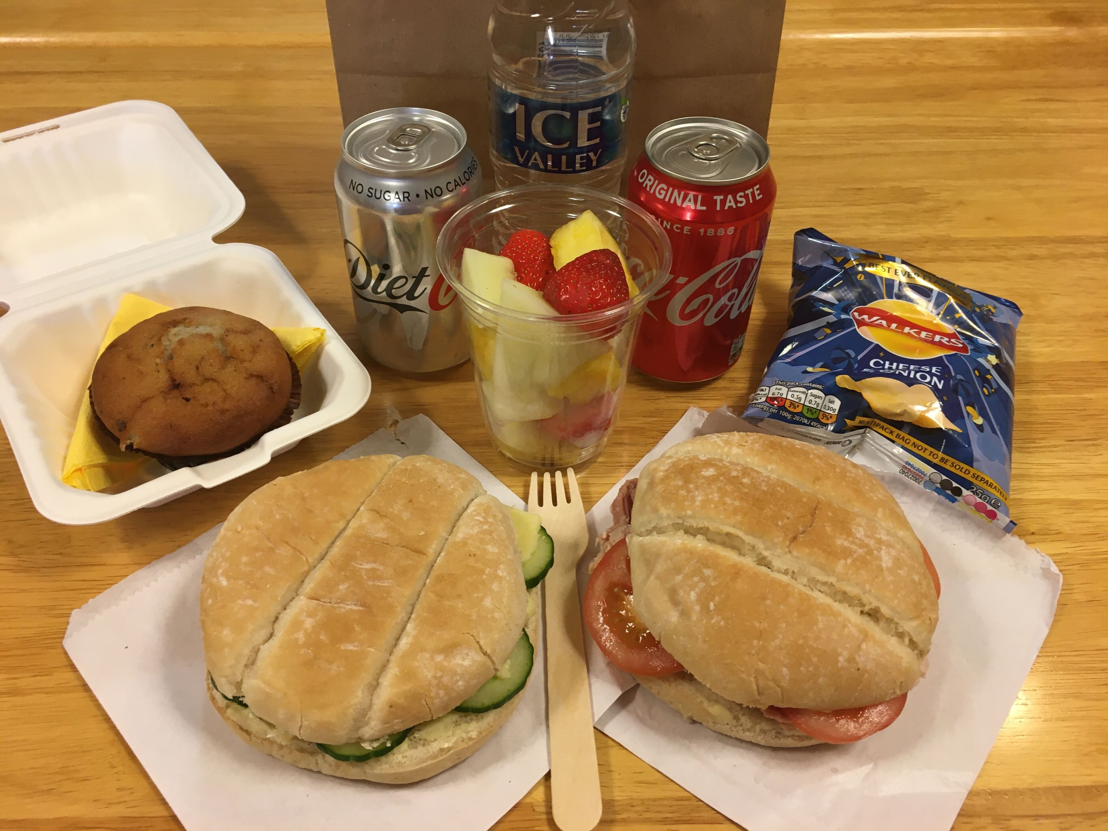 Eine Mahlzeit mit zwei Sandwiches, einem Keks, einer Obstschale und zwei Dosen Limonade auf einem Holztisch.