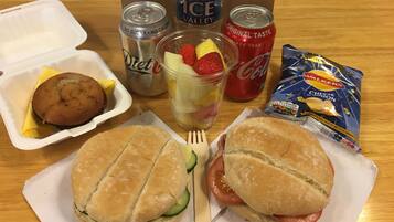 Una comida con dos sándwiches, una galleta, una ensalada de frutas y dos latas de refresco sobre una mesa de madera.