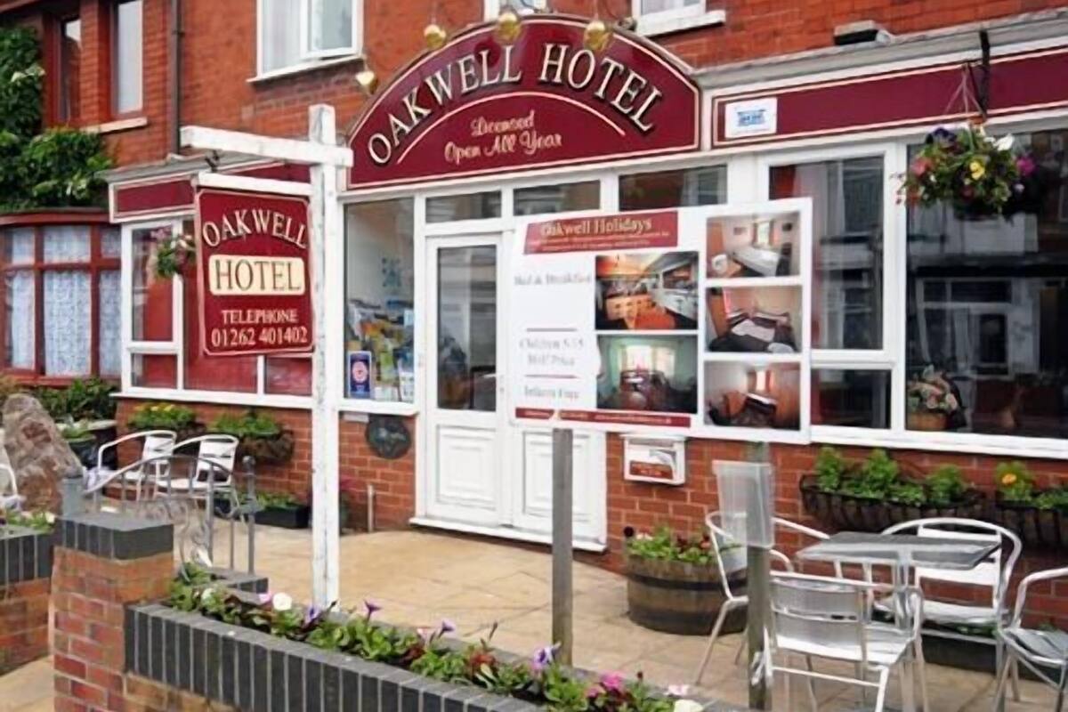 The exterior of Oakwell Hotel with a traditional facade, a visible telephone number, and a small outdoor seating area with metal chairs and tables.