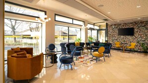 A modern hotel lobby with large windows, comfortable seating, a flat-screen TV, and a patterned wall.