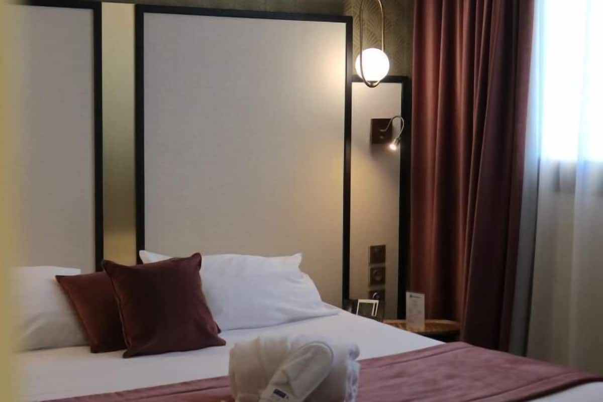 A hotel room with a bed, pillows, a bedside table, a lamp, and curtains.