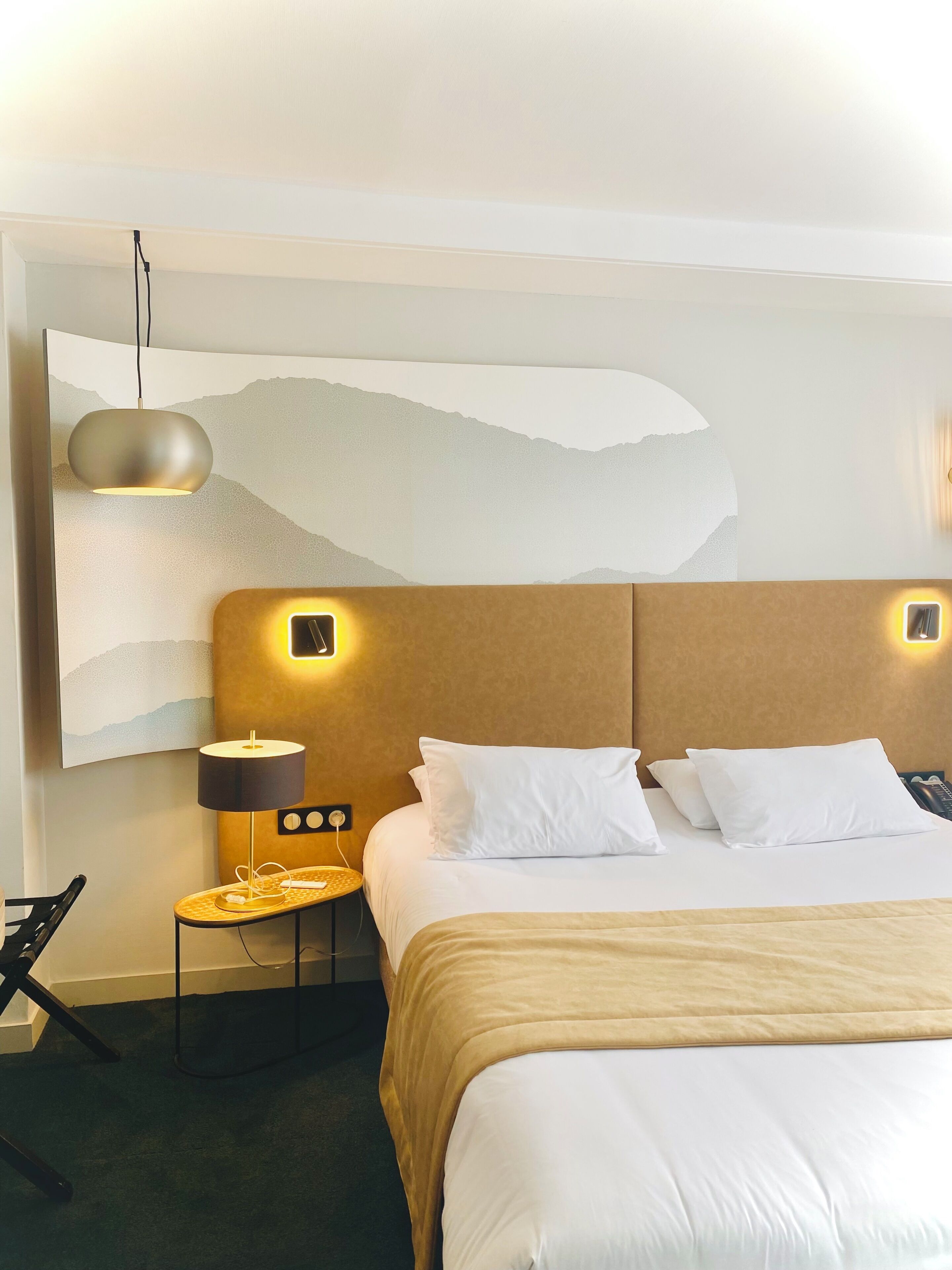 A hotel room with a large bed, a bedside table with a lamp, a wall-mounted artwork, and a modern pendant light.