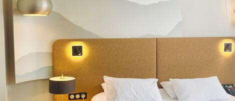 A hotel room with a large bed, a bedside table with a lamp, a wall-mounted artwork, and a modern pendant light.