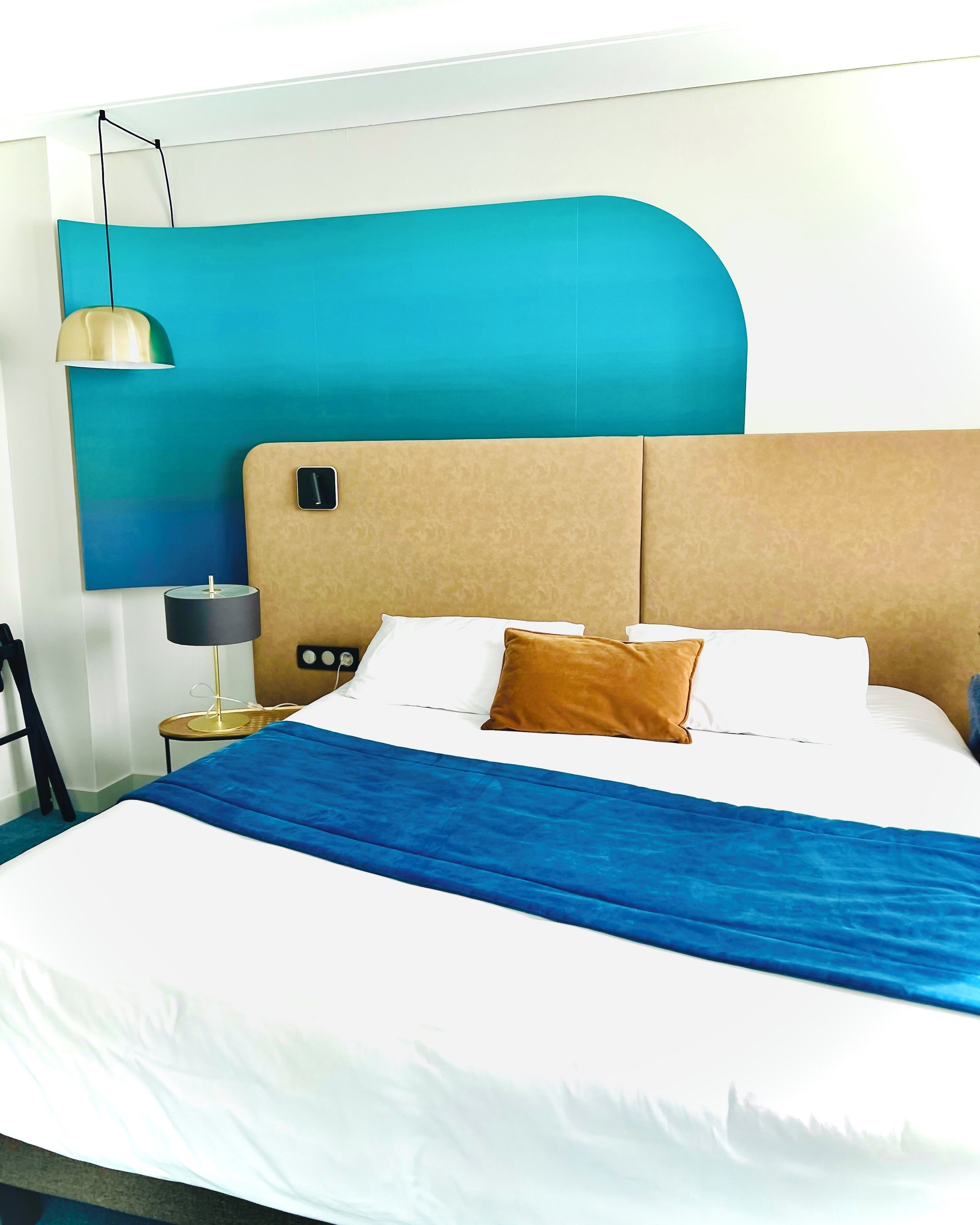 A modern hotel room with a large bed, a headboard with a control panel, a bedside table with a lamp, and a wall-mounted light fixture.