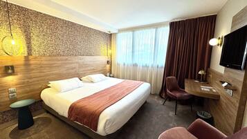 A hotel room with a bed, a desk, a chair, a television, and a wall-mounted light fixture.