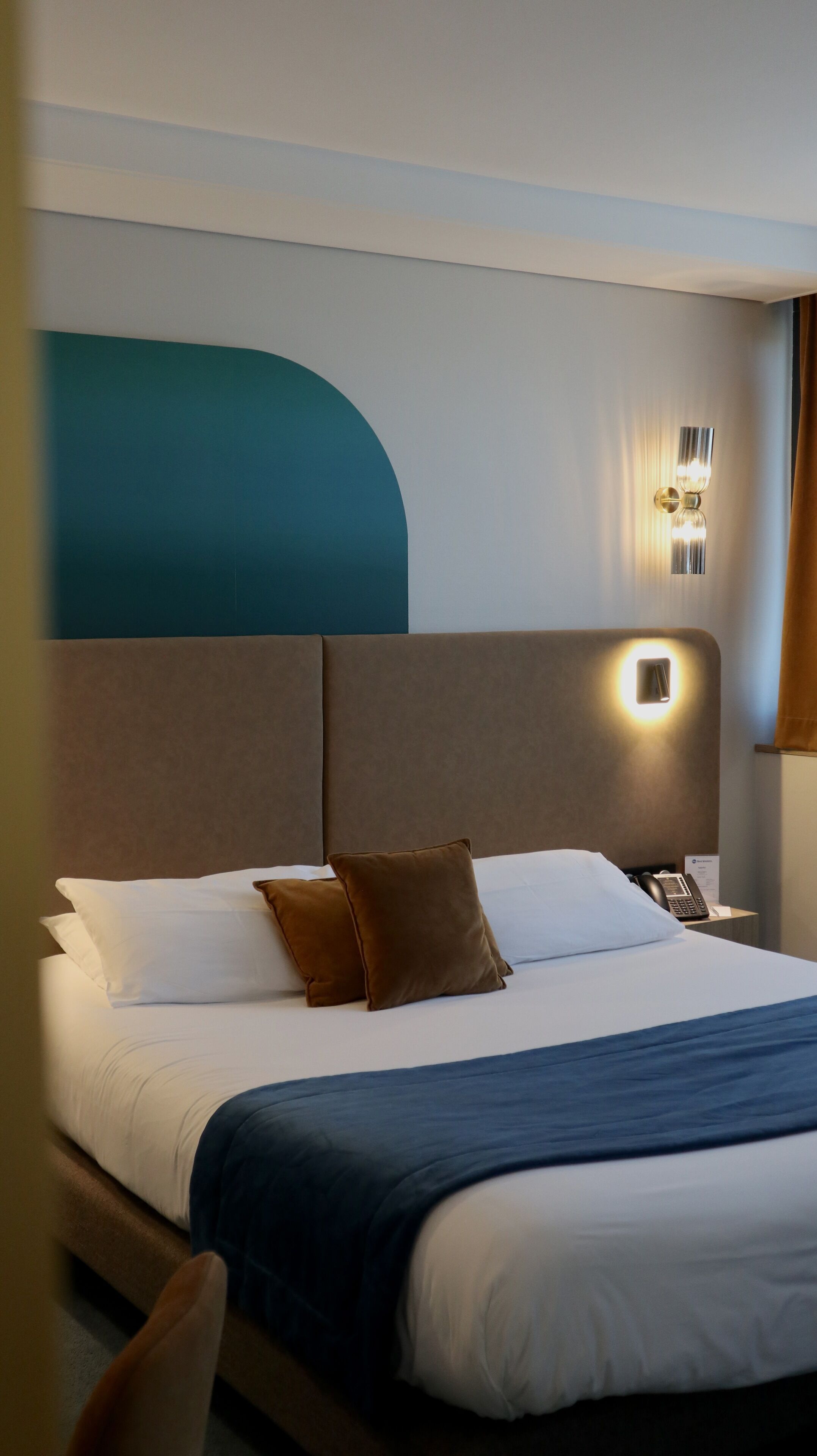 A hotel room with a bed, brown pillows, a bedside table with a phone, and a wall-mounted light fixture.