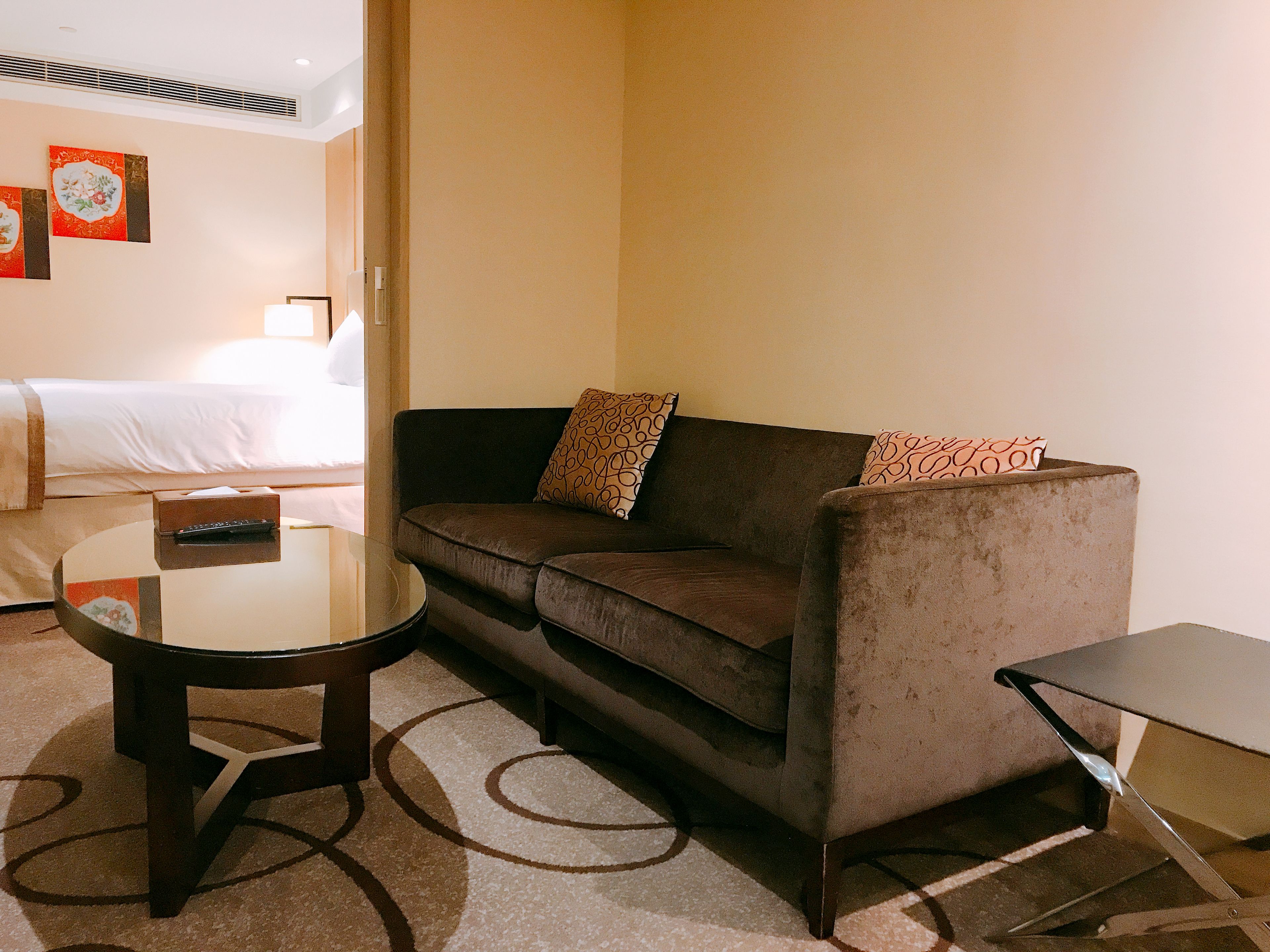 Habitación de hotel con sofá marrón, una mesita redonda pequeña, un control remoto y una cama con ropa blanca.