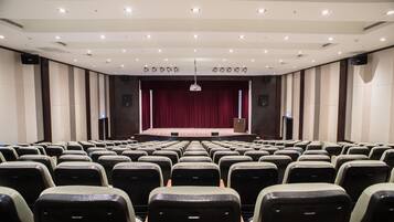 A large, empty auditorium with rows of seats, a stage with a red curtain, and ceiling lights.