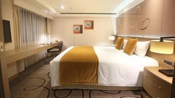 A hotel room with a large bed, a desk, a chair, and a bedside table.