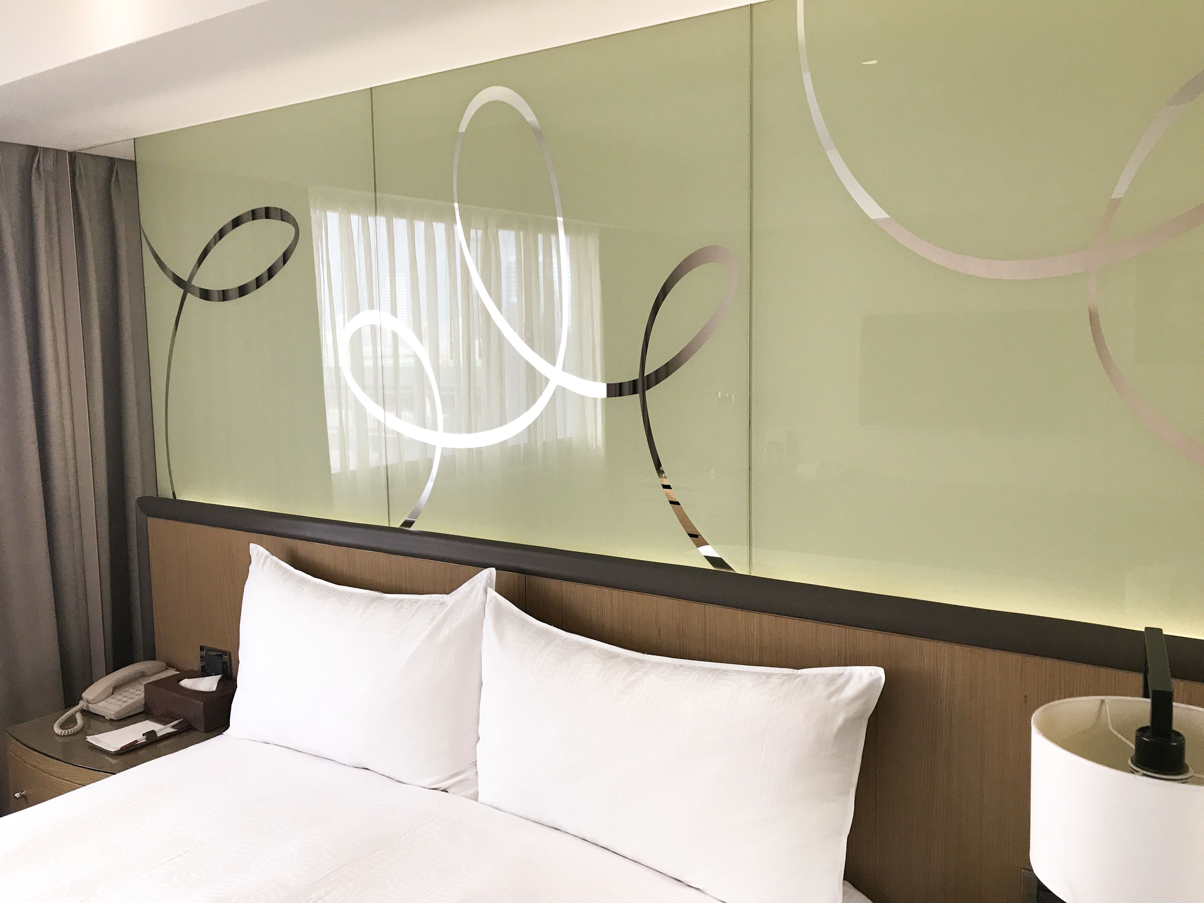 Une chambre d’hôtel avec un lit, une table de chevet avec un téléphone, une lampe et un grand miroir orné de motifs abstraits.