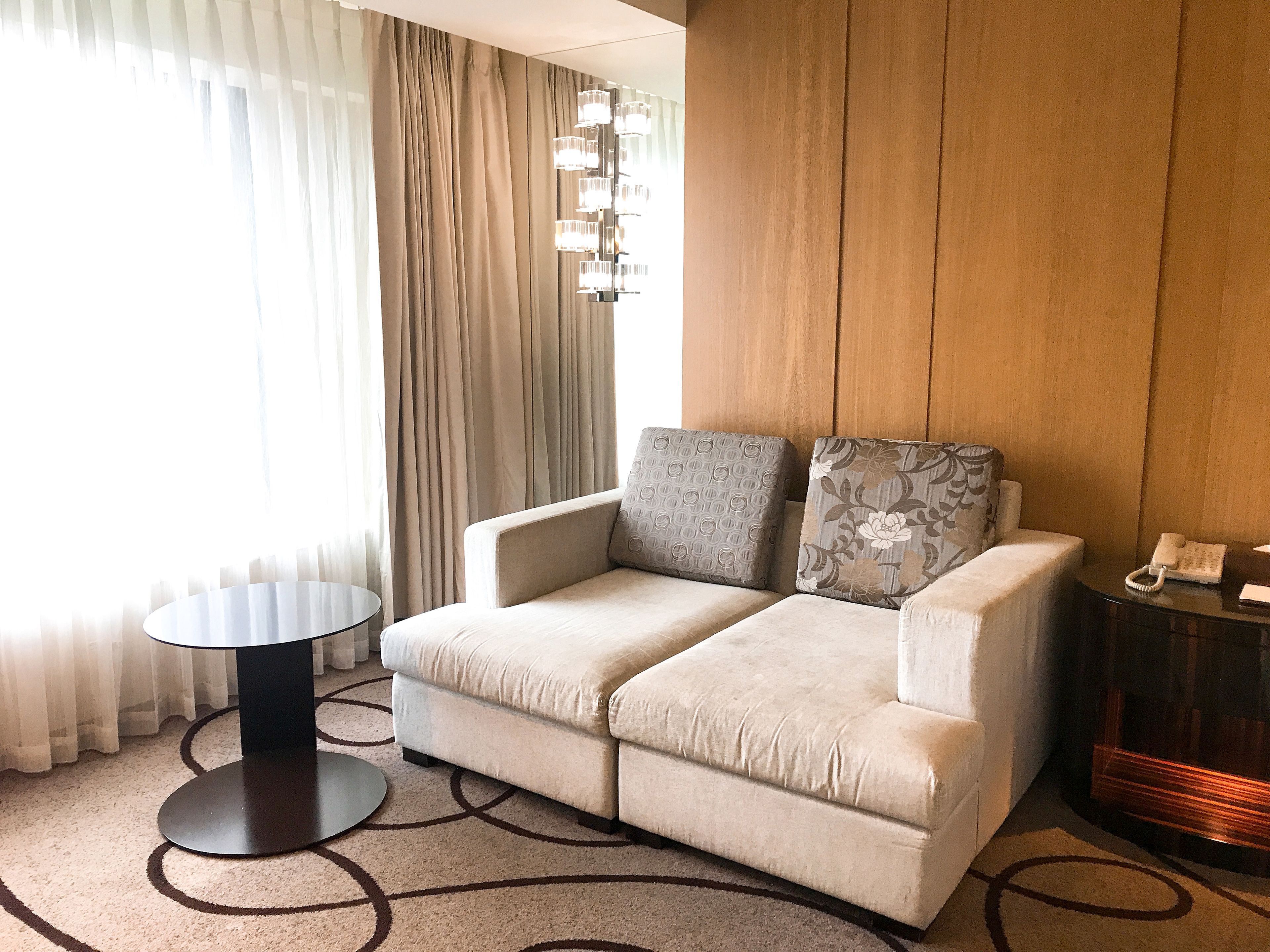 A modern hotel room with a beige sofa, two patterned cushions, a small round table, and a telephone on a wooden side table.